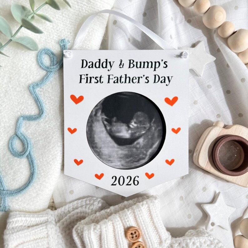 Puede incluir: Un cartel blanco con el texto "Daddy & Bump's First Father's Day" y el a&ntilde;o "2026". El cartel tiene un recorte circular que muestra una imagen de ultrasonido y est&aacute; decorado con corazones rojos.