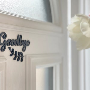 May include: A white door with a black wooden sign that says "Goodbye" with a small leafy branch underneath.