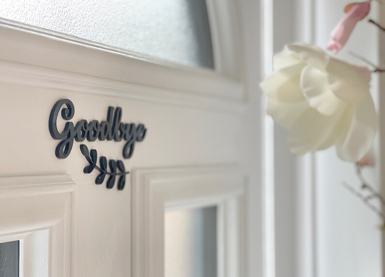 May include: A white door with a black wooden sign that says "Goodbye" with a small leafy branch underneath.