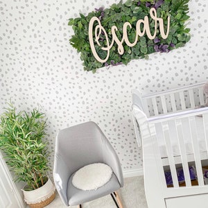 Puede incluir: Un interior de guarder&iacute;a con una cuna blanca, una silla gris con un coj&iacute;n blanco y una planta en maceta. La pared tiene un patr&oacute;n de puntos grises. Un letrero de madera con el nombre "Oscar" est&aacute; montado sobre un fondo floral verde y morado.