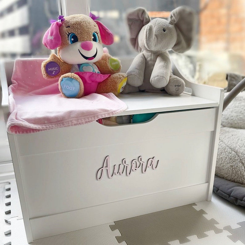 Puede incluir: Un ba&uacute;l de juguetes de madera blanca con el nombre "Aurora" en escritura cursiva. Un perro de peluche y un elefante de juguete est&aacute;n encima, junto con una manta rosa. El ba&uacute;l tiene un caj&oacute;n extra&iacute;ble y est&aacute; sobre un suelo estampado.
