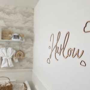 Op de afbeelding: Houten wanddecoratie met de naam Harlow geschreven in cursief schrift. De naam wordt omringd door kleine wolkvormen.
