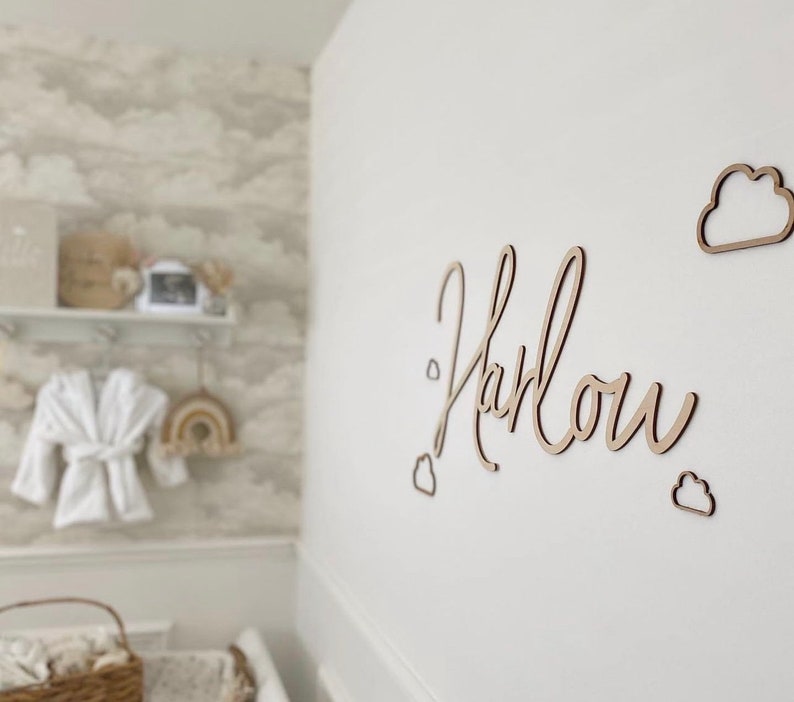 Op de afbeelding: Houten wanddecoratie met de naam Harlow geschreven in cursief schrift. De naam wordt omringd door kleine wolkvormen.