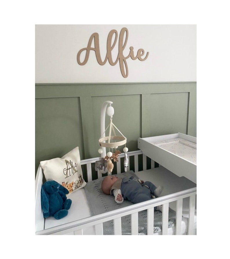 Puede incluir: Una escena de guarder&iacute;a con una cuna blanca, un m&oacute;vil y un cambiador. Un letrero de madera que dice "Alfie" est&aacute; montado en la pared. Una almohada con el nombre "Alfie" est&aacute; en la cuna. Las paredes son verdes y blancas.
