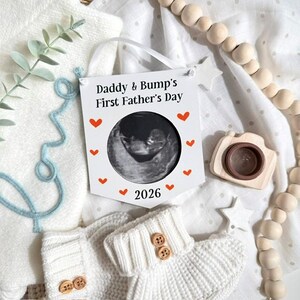 Puede incluir: Una placa blanca con el texto "Daddy & Bump's First Father's Day" y una imagen de ecograf&iacute;a. La placa est&aacute; decorada con peque&ntilde;os corazones rojos y el a&ntilde;o "2026". La imagen est&aacute; rodeada de art&iacute;culos para beb&eacute;s, incluyendo un par de botines de punto blancos.