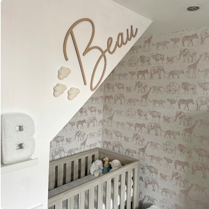 Op de afbeelding: Een wit houten babybedje met een wit en grijs behang met dierenprint. Het babybedje heeft een wit matras en een wit, pluche dekentje. Er hangen witte, pluche wolken en een houten bord met de tekst "Beau" aan de muur.