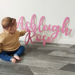 Puede incluir: Letrero de nombre de madera rosa en fuente cursiva, deletreando "Ashleigh Rose". El letrero est&aacute; apoyado contra una pared blanca. Un beb&eacute; est&aacute; sentado en el suelo, extendiendo la mano hacia el letrero.