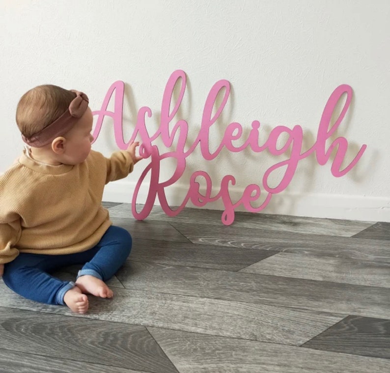 Puede incluir: Letrero de nombre de madera rosa en fuente cursiva, deletreando "Ashleigh Rose". El letrero est&aacute; apoyado contra una pared blanca. Un beb&eacute; est&aacute; sentado en el suelo, extendiendo la mano hacia el letrero.