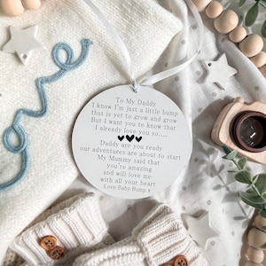 May include: A white circular ornament with a ribbon, featuring text addressed to "Daddy" from "Baby Bump." The ornament is surrounded by baby items, including a pair of cream-colored booties with wooden buttons and a blue decorative string.