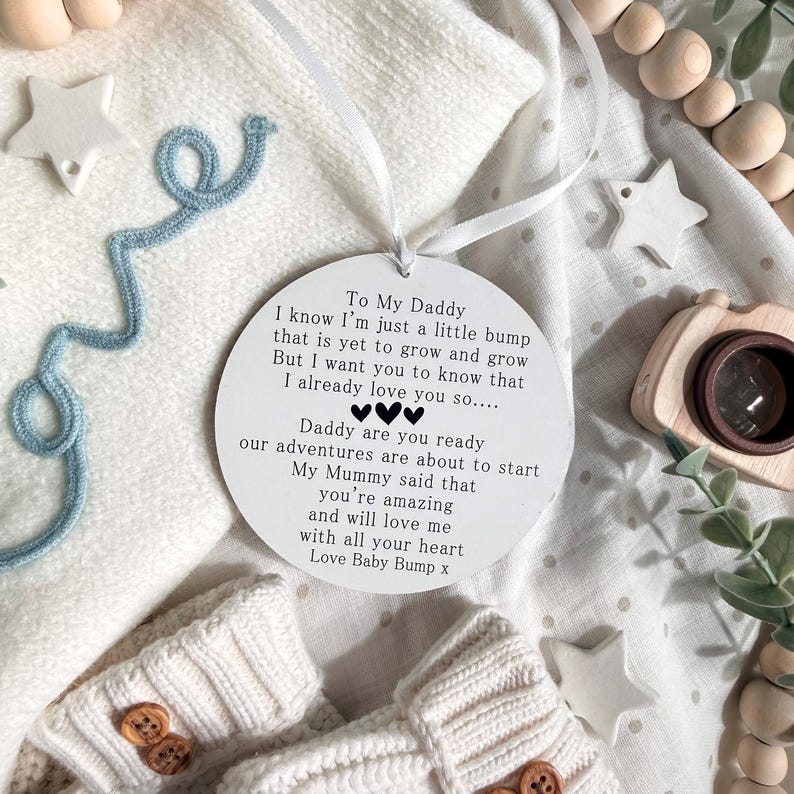 May include: A white circular ornament with a ribbon, featuring text addressed to "Daddy" from "Baby Bump." The ornament is surrounded by baby items, including a pair of cream-colored booties with wooden buttons and a blue decorative string.