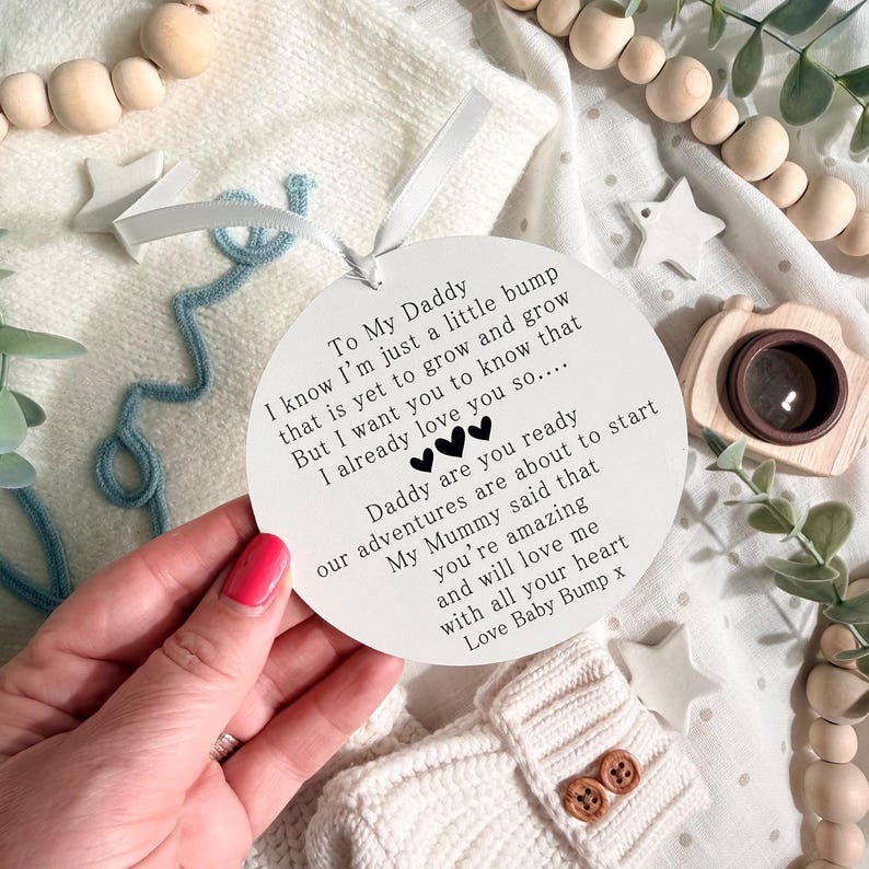 May include: A round white ornament with black text, a ribbon, and a blue arrow. The text reads "To My Daddy" and a message from "Baby Bump." The ornament is held over a white knitted baby item.
