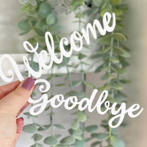 May include: White wooden cutout of the words "Welcome Goodbye" in a cursive font. The cutout is held in a hand against a background of green foliage.