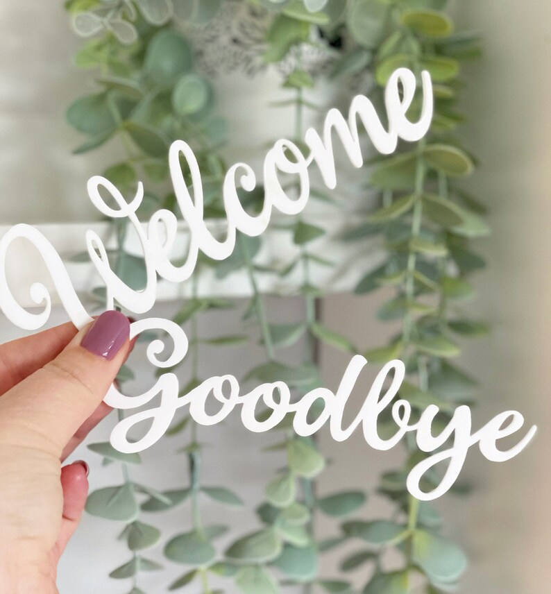 May include: White wooden cutout of the words "Welcome Goodbye" in a cursive font. The cutout is held in a hand against a background of green foliage.