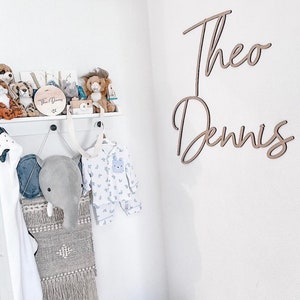 Op de afbeelding: Een witte muur met twee houten namen, "Theo" en "Dennis", geschreven in cursief schrift. Een wit rek met knuffels en een houten bord met de tekst "Theo Dennis" is aan de muur gemonteerd.