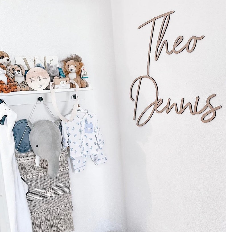 Op de afbeelding: Een witte muur met twee houten namen, "Theo" en "Dennis", geschreven in cursief schrift. Een wit rek met knuffels en een houten bord met de tekst "Theo Dennis" is aan de muur gemonteerd.