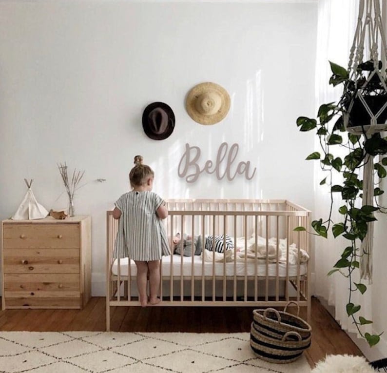Puede incluir: Una habitaci&oacute;n infantil con una cuna de madera clara, una cesta tejida y una c&oacute;moda de madera. Un ni&ntilde;o con un vestido a rayas est&aacute; de pie cerca de la cuna. El nombre "Bella" est&aacute; en la pared, con dos sombreros.