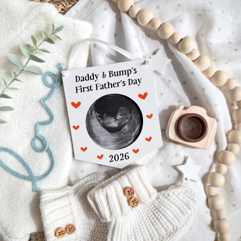 Puede incluir: Un letrero blanco con el texto "Daddy & Bump's First Father's Day" y una imagen de ultrasonido. Acentos de coraz&oacute;n rojo rodean el ultrasonido y el a&ntilde;o "2026". La imagen incluye botines de beb&eacute;, una c&aacute;mara de madera y una guirnalda de cuentas de madera.