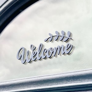 May include: A grey welcome sign with a stylised font and a small leaf design. The sign is attached to a glass door.