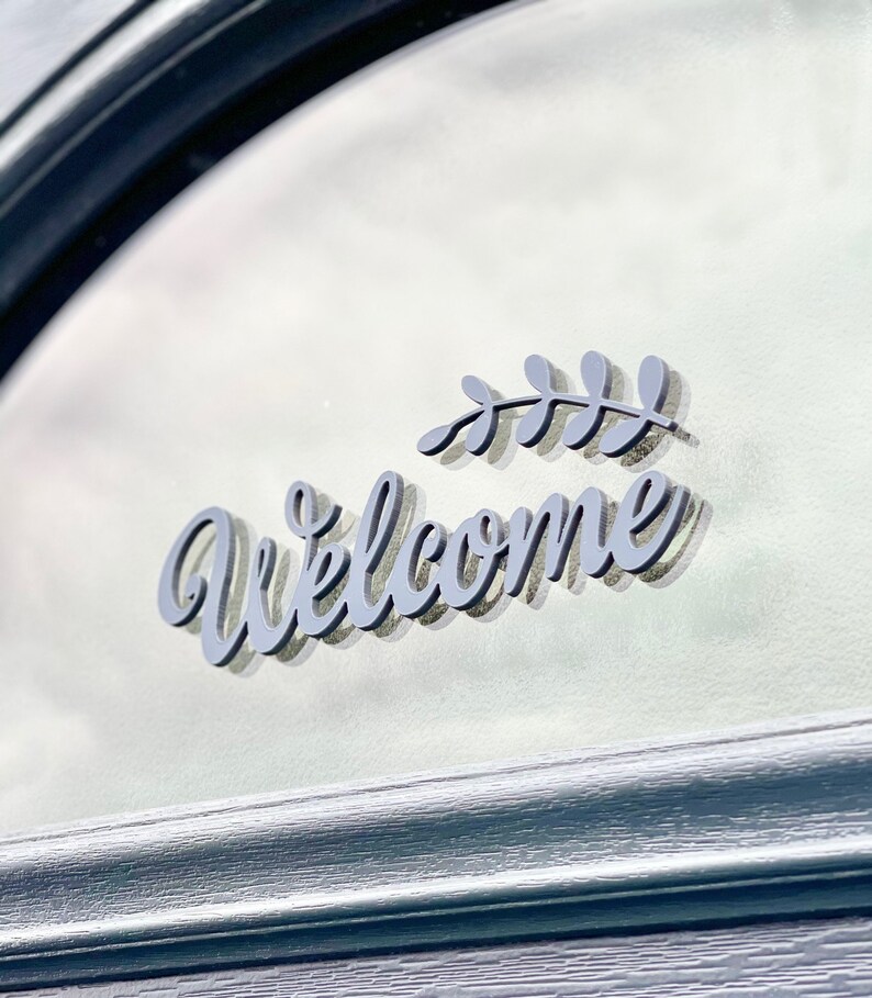 May include: A grey welcome sign with a stylised font and a small leaf design. The sign is attached to a glass door.