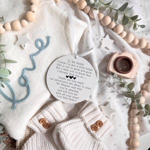 May include: A white, round ornament with text, a pair of baby booties, and wooden beads. The ornament has a ribbon for hanging and reads "To My Daddy" with a loving message. The booties are cream-colored with wooden buttons.