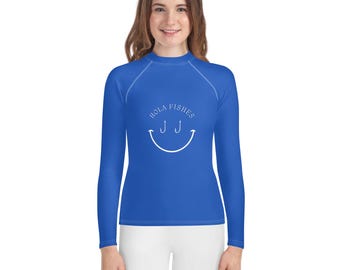 May include: A royal blue long-sleeved rash guard with a white smiley face design. The smiley face has two fishing hooks for eyes and the text "Hola Fishes" above it.