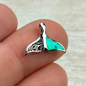 Könnte beinhalten: Ein kleiner, silberfarbener Anhänger in Form einer Walflosse. Die Flosse hat auf der einen Seite aufwendige filigrane Aussparungen und auf der anderen Seite eine leuchtend türkisfarbene Einlage. Der Anhänger ist als Schmuckstück konzipiert.