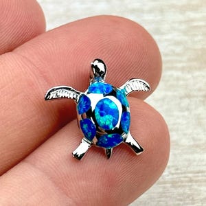 Op de afbeelding: Een zilverkleurige schildpendant met een blauwe opaal inleg. De hanger heeft gedetailleerde vin- en hoofdontwerpen. De opaal heeft een mix van lichte en donkere blauwtinten. Een klein sieraad.