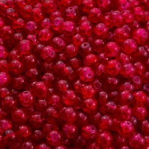 Puede incluir: Un cuenco blanco con borde festoneado lleno de numerosas cuentas rojas translúcidas pequeñas. Las cuentas están densamente empaquetadas, creando una superficie vibrante y texturizada. Las cuentas parecen estar hechas de vidrio o un material similar, reflejando la luz.