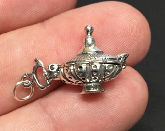 Puede incluir: Un amuleto de plata en forma de lámpara de genio adornada con una tapa con bisagras. El amuleto tiene un bucle para sujetarlo a un collar o pulsera.
