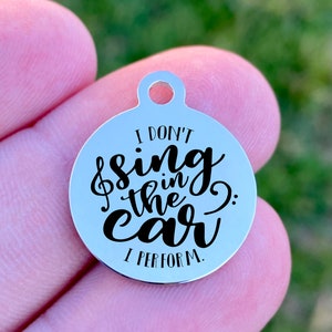 May include: A round, silver-colored charm with a hole at the top. The charm has the text "I DON'T sing in the car I PERFORM" in black script. The charm is held in a person's hand.