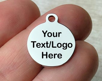 May include: A small, circular, silver-coloured charm with a hole at the top. The charm features the text "Your Text/Logo Here" in black, centrally placed. The charm is held in a person's hand.