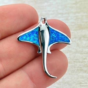 Puede incluir: Un colgante plateado con forma de manta raya. Las alas están incrustadas con un material azul brillante y moteado. El colgante se sostiene en una mano, destacando el diseño detallado y el tema marino.