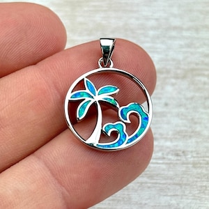 May include: A silver pendant with a tropical theme. The pendant features a palm tree and ocean wave design, with turquoise and blue accents. The pendant is round and has a small loop at the top for a chain.