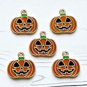 May include: Five orange and black enamel pumpkin charms with gold-colored trim. Each charm features a jack-o'-lantern face with a green stem and a loop for attaching to jewelry or other crafts. The charms are arranged on a white wooden surface.