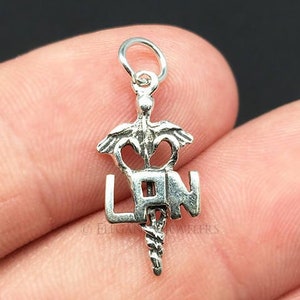 May include: Silver charm with a medical caduceus symbol and the letters "RN" in the center. The charm is on a finger.