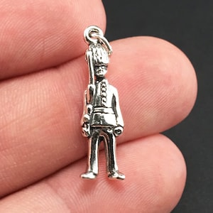 May include: Silver charm of a standing soldier wearing a tall hat and holding a rifle. The charm is about 2 inches tall.