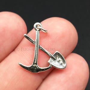 Puede incluir: Encanto de plata con un ancla y una pala cruzados, con un bucle para sujetarlo a un collar o pulsera.