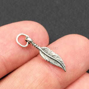 May include: A small, silver-colored feather charm with detailed veining. The charm has a loop at the top for attaching to a necklace or bracelet. The feather is held between fingers against a dark background. The text "Elegancy Jewelers" is at the bottom.