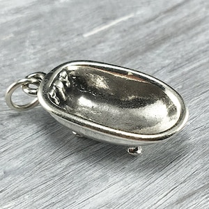 Puede incluir: Un colgante plateado con forma de bañera. La bañera en miniatura tiene un interior detallado y se apoya sobre pequeñas patas. Se adjunta un lazo para una cadena. El colgante es una joya.