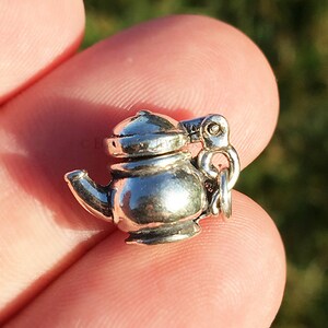 May include: A silver charm in the shape of a teapot. The charm has a detailed design and a small loop for attaching to a bracelet or necklace.