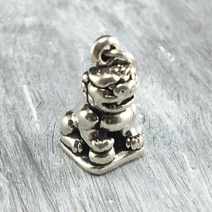 May include: A small, silver-colored charm in the shape of a stylized lion-like creature. The charm has detailed features, including a textured mane, a smiling face, and is sitting on a square base. The charm has a loop at the top for attachment. The text "Elegancy Jewelers" is at the bottom.