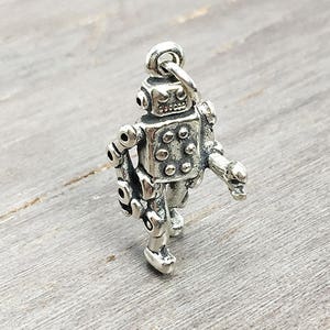 May include: A silver charm in the shape of a robot with a round head and a body with gears and bolts. The robot is walking with one arm raised.