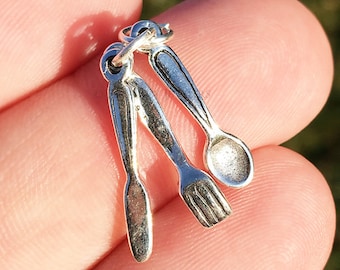 Może przedstawiać: Srebrny charm z zestawem trzech miniaturowych przyborów kuchennych: widelcem, nożem i łyżką. Charm jest zaprojektowany do przymocowania do bransoletki lub naszyjnika.