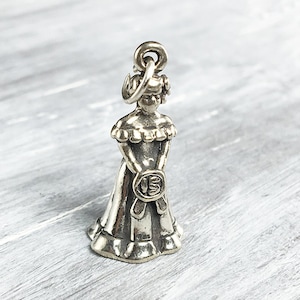 May include: Silver charm of a girl in a dress holding a '15' in her hands. The charm is a popular choice for quinceañera celebrations.
