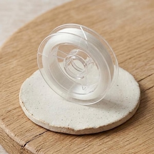 May include: A clear, transparent spool of beading thread sits on a small, off-white ceramic disc. The disc rests on a wooden surface. The thread is tightly wound within a clear plastic spool, ready for jewelry making or crafts.