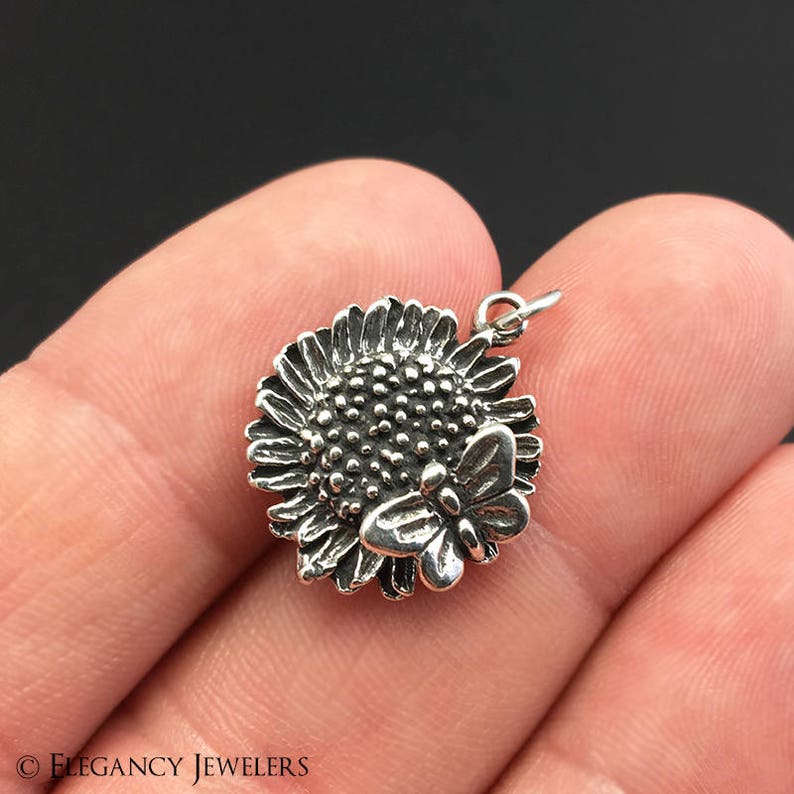May include: A silver charm featuring a detailed sunflower with a butterfly perched on top. The charm has a loop for attaching to a necklace or bracelet.