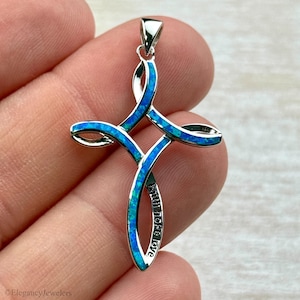 Puede incluir: Un colgante plateado en forma de cruz con incrustaciones de ópalo azul. El diseño de la cruz presenta un patrón entrelazado con las palabras "Faith Hope Love" grabadas en la parte inferior. El colgante tiene un pequeño lazo en la parte superior para una cadena.
