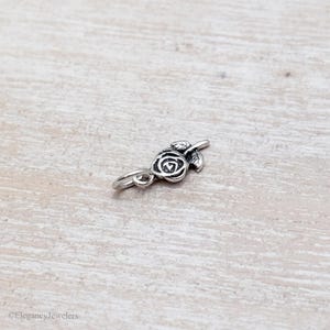 May include: A small silver-toned metal charm shaped like a rose blossom with a single leaf and an attached jump ring. The surface features oxidized detailing to highlight the floral contours and the text ElegancyJewelers is visible at the bottom.