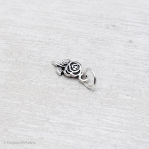 May include: A small silver-toned metal charm shaped like a rose flower with detailed petals and leaves. The charm features an oxidized finish to highlight the engraved floral design and includes a circular jump ring attached to the stem for hanging.