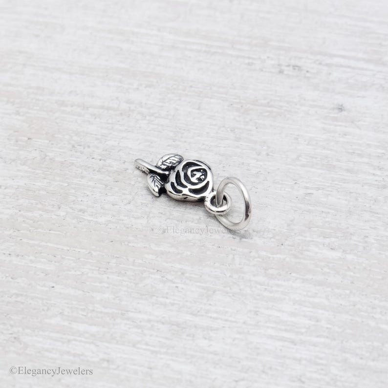 May include: A small silver-toned metal charm shaped like a rose flower with detailed petals and leaves. The charm features an oxidized finish to highlight the engraved floral design and includes a circular jump ring attached to the stem for hanging.